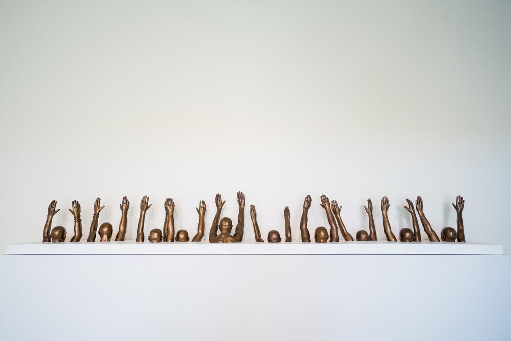 Gold sculptures of heads and arms raised in the air are shown in front of a white space