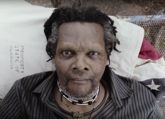 Video still of a black man with a lot of necklaces lying in a bed staring directly at the camera. From a Lonnie Holley video.