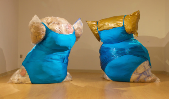 two large trash bags filled with smaller, crumpled plastic bags. the trash bags are filled to the seems and wear turquoise blue spandex dresses, so that the filled trash bag bulges out of the dress like human skin