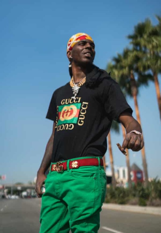 Young Dolph smiling carefree on the side of a street lined with tall palm trees. He wears bright green pants with a red and gold gucci belt, a black Gucci shirt with the same colors, and a red and gold durag. He is iced out with jewelry and a gold watch, he holds a blunt in his left hand and looks at something in the distance.