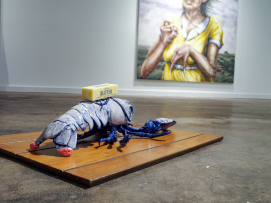 installation of a large blue lobster statue with salted butter on its back, the lobster is on a brown wooden board with thick panels, a painting of a figure in a yellow dress hangs on a wall in the background