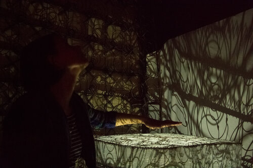 a shadowed figure holds a hand over a covered surface, winding shadows dance in the space, cast from the bed springs on the adjacent wall, the photo looks like a seance