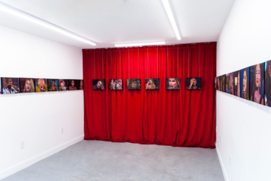 white gallery walls with a crimson red curtain spanning the center back wall. in a line spanning the three walls are painted portraits of femmes crying