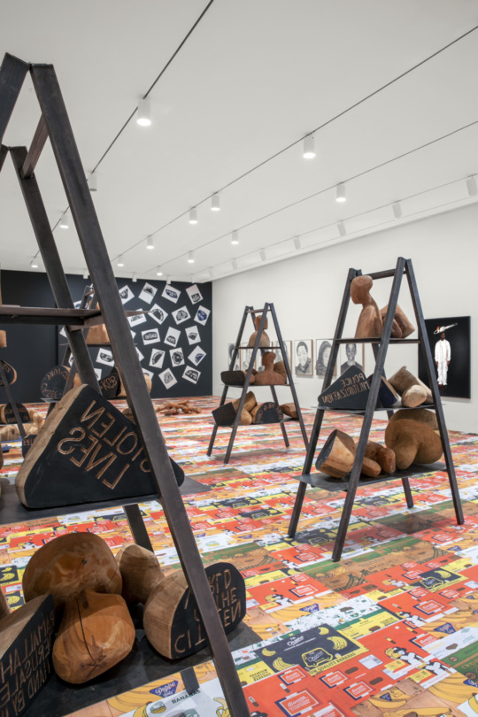 large ladders in a white and black walled gallery space with colorful flooring. on the ladders are large stone-like sculpture with wooden accents with phrases like 