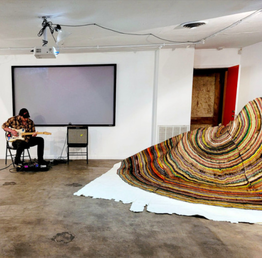 a person sits and performs a soundcheck alongside a large painting work on white canvas