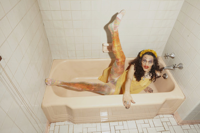 film photograph of a drag queen wearing a yellow outfit with colorful tights inside of a white bathroom in a light pink tub
