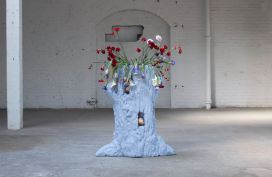 a white sculpted tree trunk stump with flowers spilling out the top in a warehouse gallery space