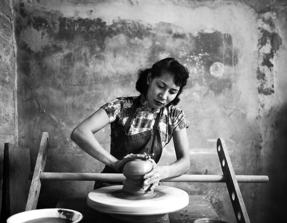 A woman sits center of the black and white photograph, with an apron on and uses both hands to mold a piece of clay on a pottery wheel.