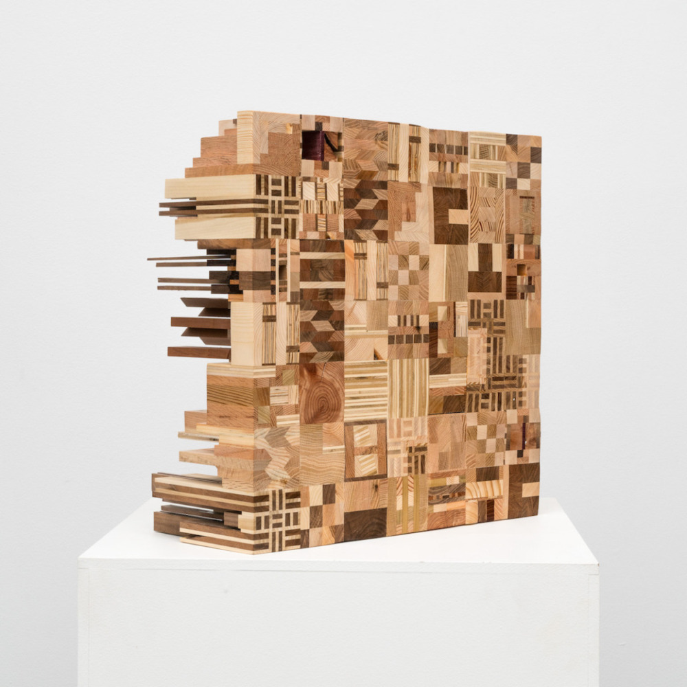 a large wooden block sits on a white cube. built into the wooden block are many different shapes, made with different shades of wood. 