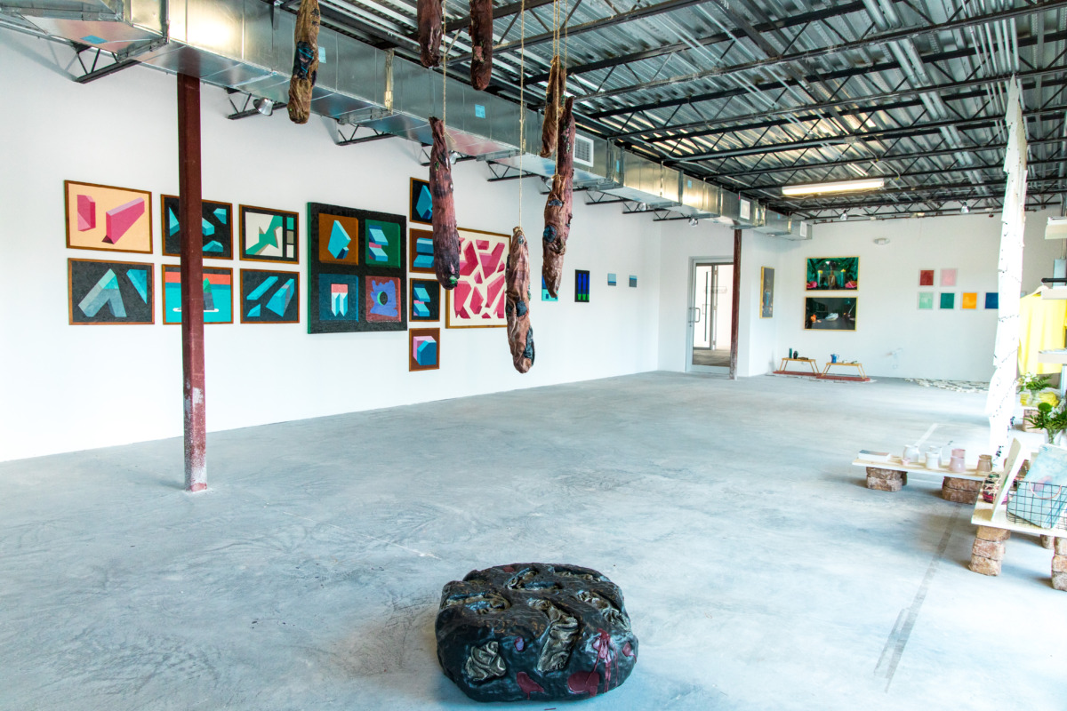 An installation show of a large warehouse space with white walls, steel ceiling, and concrete floor. Paintings of shapes on the white wall, hanging textiles from the ceiling and a large black stone like object on the floor in the foreground.