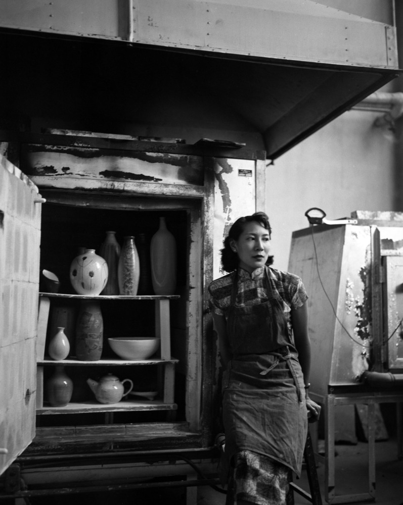 A black and white photograph of a woman with an apron on stands against a kiln looking to the side. The kiln is filled from top to bottom with ceramic pieces.