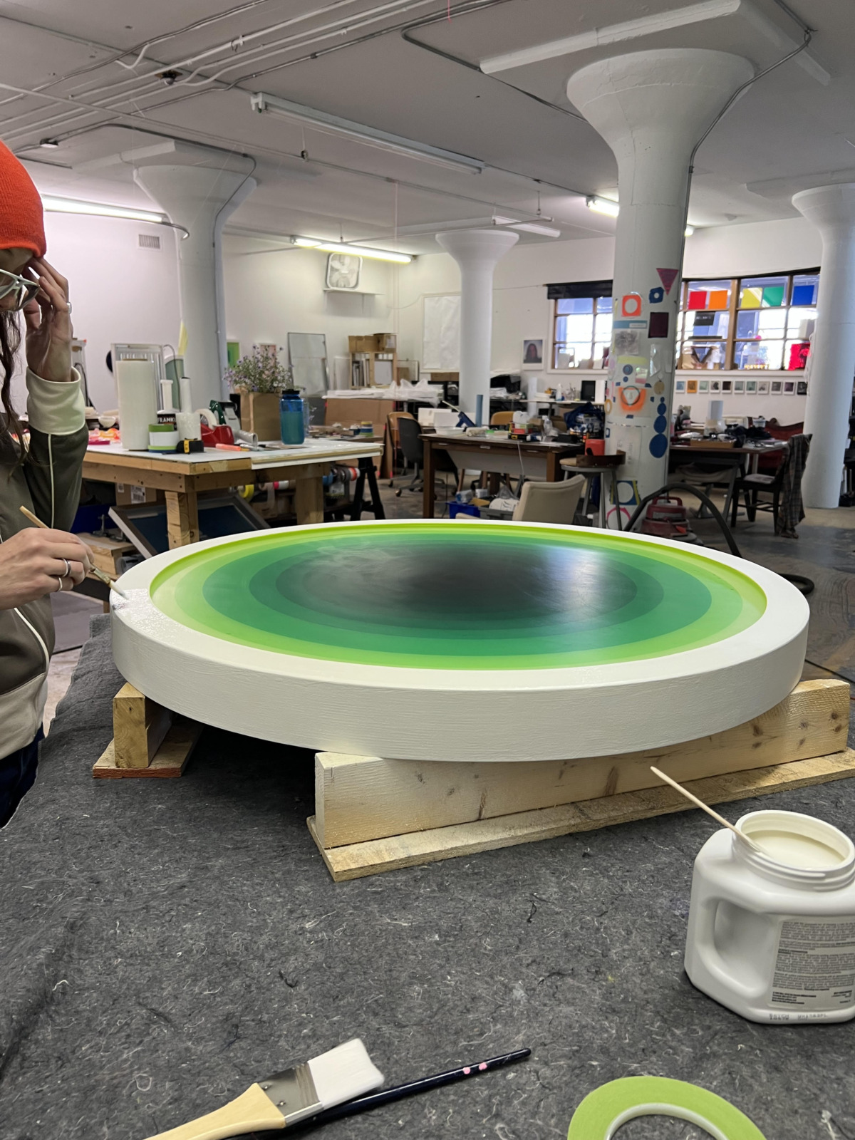 a busy studio with large white pillars, with tables covered in art supplies. in the foreground, a white woman with an orange beanie and rings on her hands works on a large circular painting with reverberating shades of green