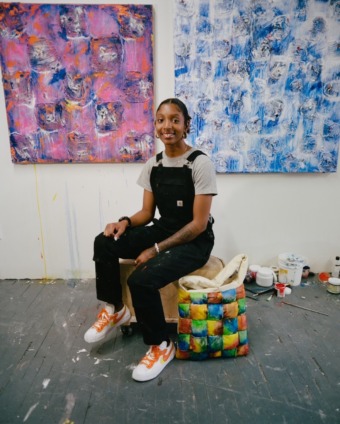 a photo of an artist wearing black overalls and a white t-shirt. they sit in front of two colorful paintings on a white wall in an industrial gallery space