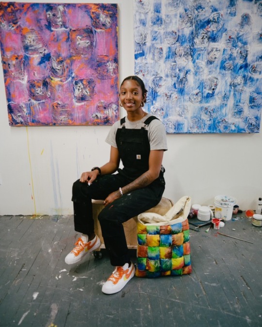 a photo of an artist wearing black overalls and a white t-shirt. they sit in front of two colorful paintings on a white wall in an industrial gallery space