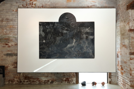 Installation image of white hanging square with black and white etching on top, depicts ambiguous figures in a wilderness scene under the night sky.