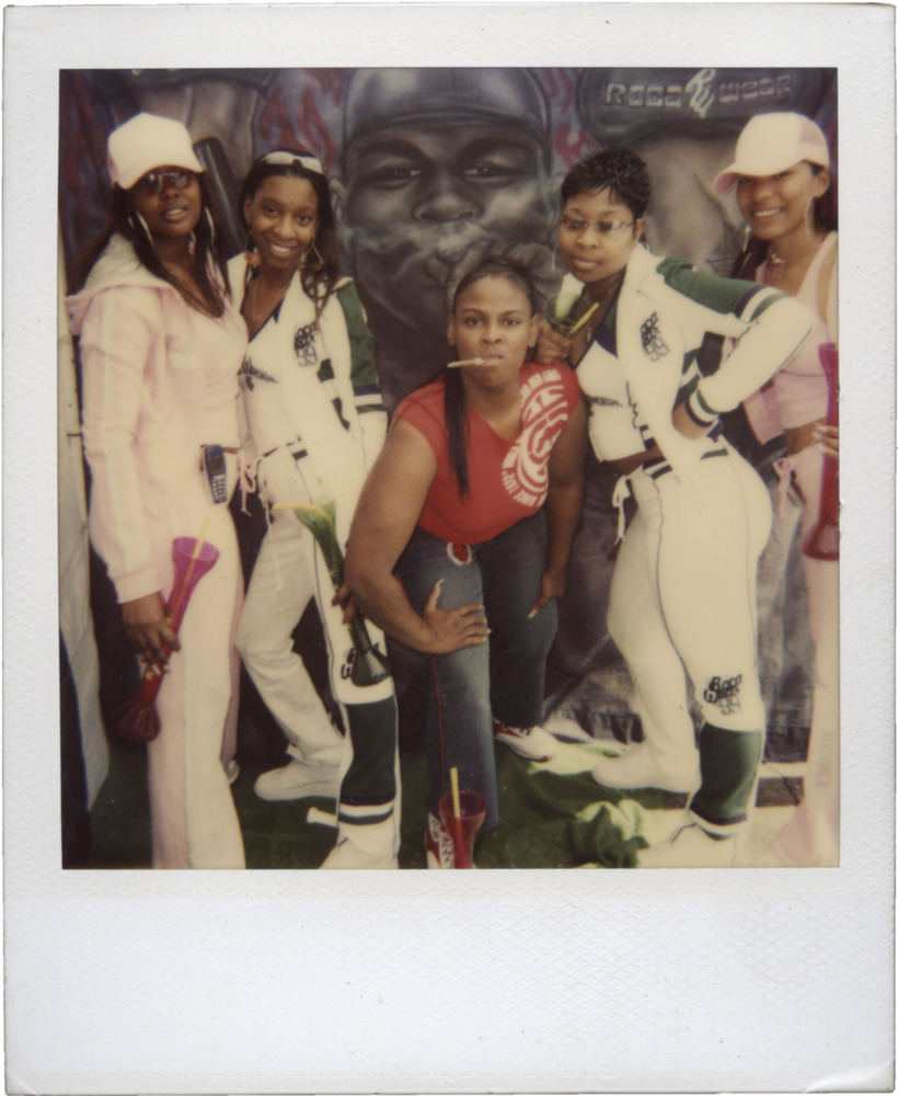 A polaroid shows a group of five black women posing in front of a graffiti backdrop with the woman in the center crouching forward with a lollipop in her mouth