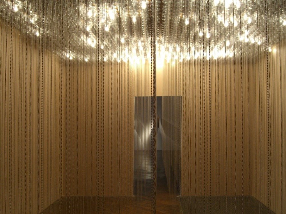 a golden-lit exhibition space filled with hanging metal chains and flickering lights